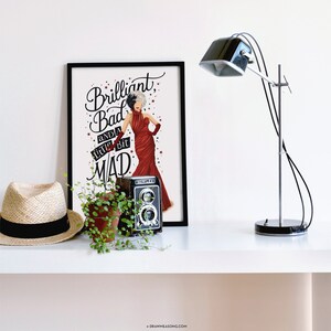 May include: A black framed print with a white background featuring a cartoon illustration of Cruella de Vil in a red dress with the text "Brilliant, Bad and a little bit Mad".