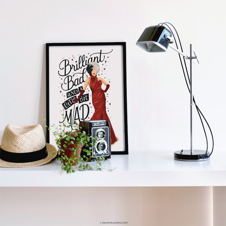 May include: A black framed print with a white background featuring a cartoon illustration of Cruella de Vil in a red dress with the text "Brilliant, Bad and a little bit Mad".