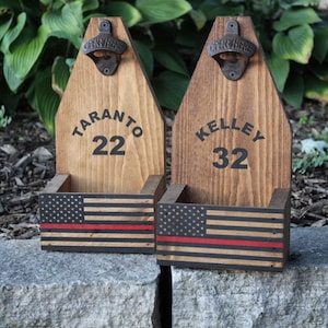 May include: Two wooden bottle openers with a red, white, and blue American flag design. The bottle openers have a metal bottle opener attached to the top and a small shelf below for holding bottles. The top of each bottle opener is personalized with a name and number. The first bottle opener says "TARANTO 22" and the second bottle opener says "KELLEY 32".