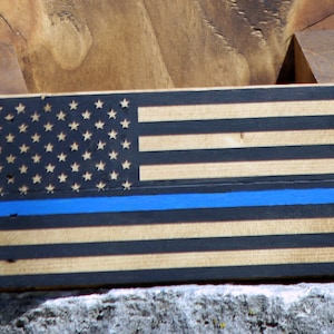 May include: Wooden bottle opener with a thin blue line American flag design. The flag is black, white, and brown with a blue line across the middle.