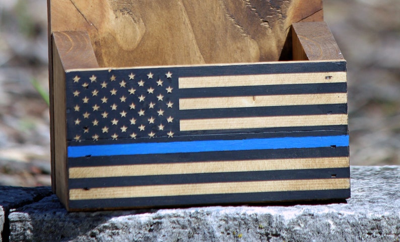 May include: Wooden bottle opener with a thin blue line American flag design. The flag is black, white, and brown with a blue line across the middle.