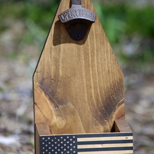 May include: Wooden bottle opener with a thin blue line American flag design. The bottle opener has a metal opener with the words "Open Here" on it.