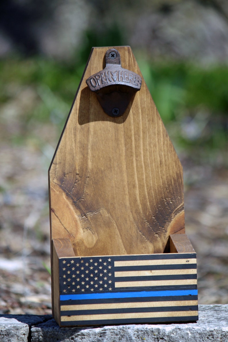 May include: Wooden bottle opener with a thin blue line American flag design. The bottle opener has a metal opener with the words "Open Here" on it.