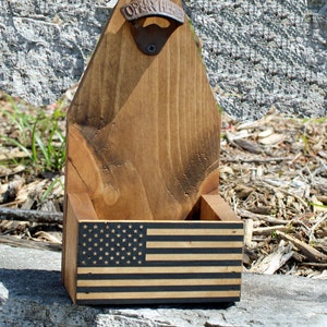 May include: Wooden bottle opener with a built-in bottle holder. The bottle opener has a metal "Open Here" opener and a black and white American flag design on the front.
