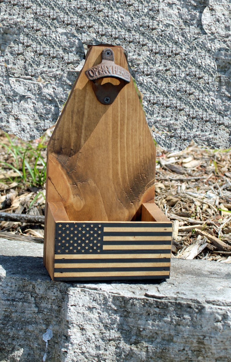 May include: Wooden bottle opener with a built-in bottle holder. The bottle opener has a metal "Open Here" opener and a black and white American flag design on the front.
