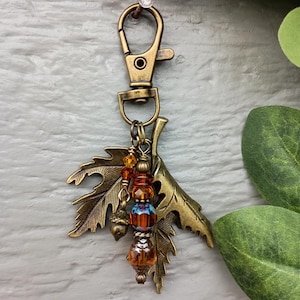May include: A bronze-toned metal keyring featuring a lobster clasp and two maple leaf charms. A vertical strand of beads hangs between the leaves, including amber-coloured faceted crystals, a small metal acorn, and a blue and orange patterned bead.