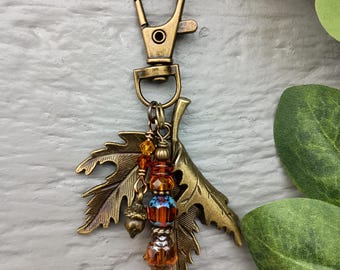 Op de afbeelding: Een bronzen metalen sleutelhanger met een karabijnhaak en twee esdoornbladbedels. Tussen de bladeren hangt een verticaal snoer van kralen, waaronder barnsteenkleurige gefacetteerde kristallen, een kleine metalen eikel en een blauwe en oranje kraal.