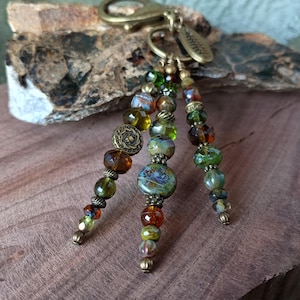 May include: A beaded keychain with a brass clasp and decorative charms. The keychain features strands of colorful beads in shades of green, brown, and blue. The beads vary in size and shape, with some having a textured appearance. The keychain is displayed on a wooden surface.