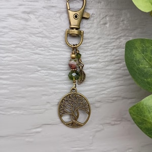May include: A bronze-colored keychain with a tree of life pendant. The keychain features a lobster clasp, beads in shades of green and red, and a small leaf charm. The tree pendant is a detailed cutout design.