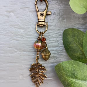 May include: A bronze-toned metal lobster clasp keychain featuring a dangling charm set. The set includes a faceted iridescent glass bead, a small bronze acorn charm, and a detailed bronze leaf charm, all connected by wire-wrapped metal rings.