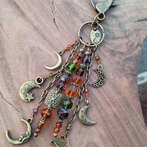 May include: A bronze-toned keychain featuring multiple dangling chains, faceted glass beads in amber, green, and purple, and various moon and star charms. The accessory includes a circular ring and a lobster clasp for attachment to keys or bags.