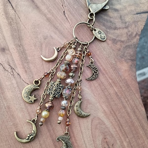 May include: A bronze-toned keychain featuring a lobster clasp and a circular ring. Multiple dangling chains are adorned with various moon and star charms, along with faceted glass beads in shades of amber, brown, and clear, creating a layered look.