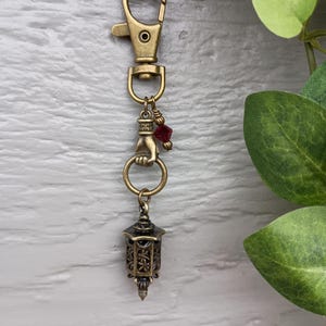 Op de afbeelding: Een messingkleurige metalen sleutelhanger met een karabijnhaak, een handvormige bedel die een ring vasthoudt, een kleine gefacetteerde rode kraal en een miniatuur lantaarnhanger met gedetailleerde uitgesneden patronen.