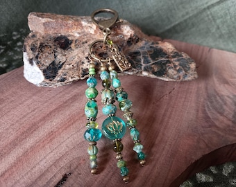 May include: A beaded keychain with turquoise, green, and gold accents. The keychain features multiple strands of beads in varying sizes and shapes, with a central charm displaying a lotus flower design. The metal hardware has an antique brass finish.