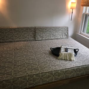 May include: A large, rectangular bed with a patterned fabric cover. The bed has a wooden frame and is sitting on a carpeted floor. A small tray with a throw blanket and a remote control is on the bed.