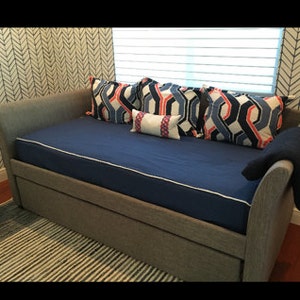 May include: A gray upholstered daybed with a blue mattress and a white trim. The daybed has a geometric patterned throw pillow in blue, red, and white. There is a white throw pillow with red and blue accents. The daybed is in front of a white and gray chevron patterned wall.