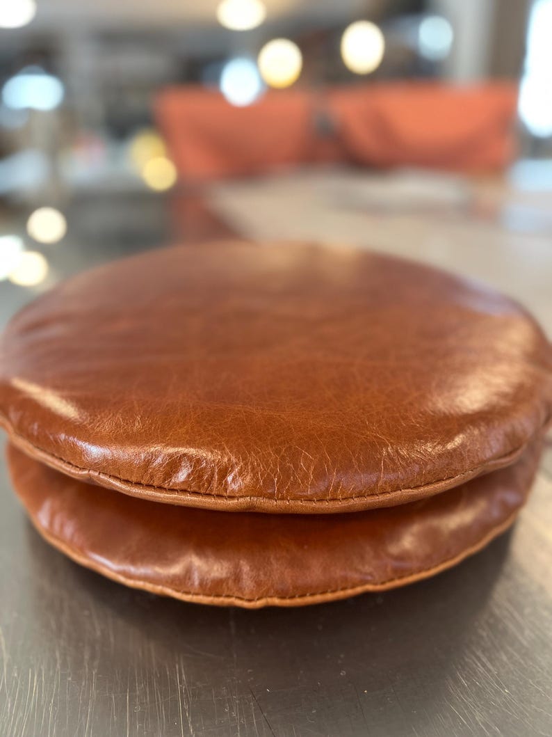 May include: Two brown leather round cushions stacked on top of each other. The cushions are stitched together with brown thread.