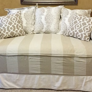 May include: A white and beige striped daybed with a white skirt. The daybed has five pillows, two are beige with a geometric pattern, two are white with a floral pattern, and one is beige with a paisley pattern.