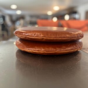 May include: Two brown leather cushions stacked on top of each other. The cushions are round and have a smooth, slightly textured surface.