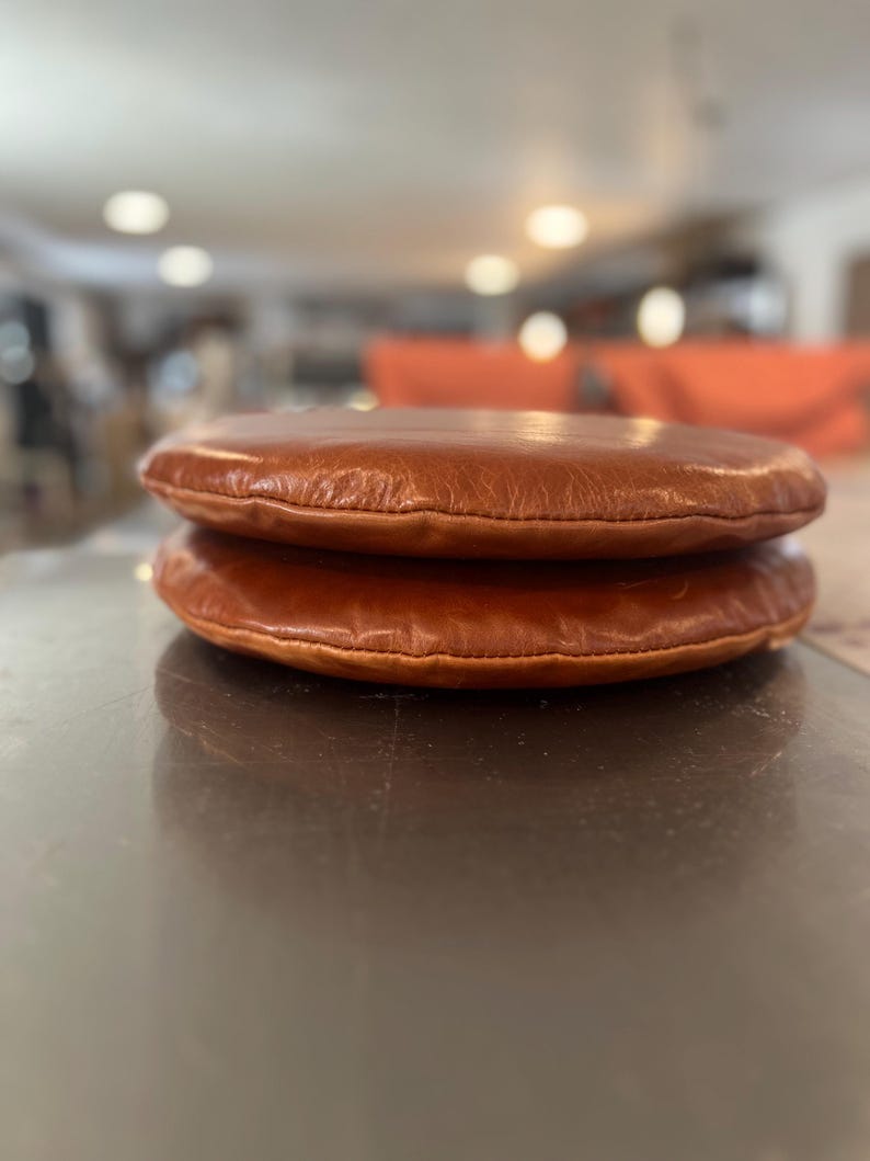 May include: Two brown leather cushions stacked on top of each other. The cushions are round and have a smooth, slightly textured surface.