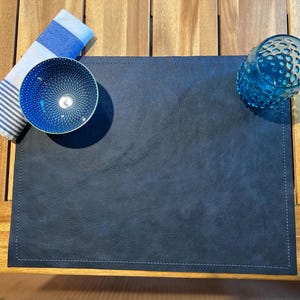 May include: A navy blue placemat with white stitching, a blue patterned bowl, a blue glass, and a blue and white striped napkin on a wooden table. The placemat is rectangular and has a textured surface.