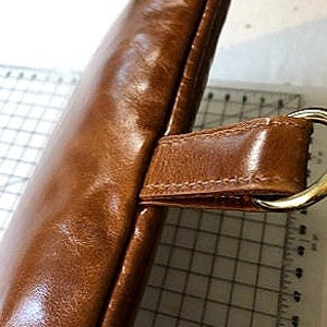 May include: Close-up of a brown leather bag with a gold metal ring and a strap. The bag has a smooth, glossy finish.