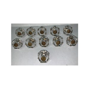 May include: A collection of eleven clear, faceted, octagonal glass knobs. Each knob features a central, circular, metallic gold-toned decorative insert with a textured, floral-like pattern on the front face, arranged in a grid-like formation.