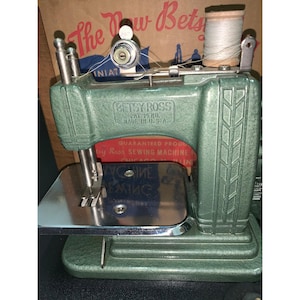 May include: A vintage green Betsy Ross sewing machine with a metallic finish, featuring a chrome needle plate and a spool of white thread on top. The machine body is embossed with the text Betsy Ross Pat. Pend. Made in U.S.A. and sits before its box.