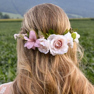 Puede incluir: Una corona floral con lirios rosa claro, rosas rubor y flores blancas con hojas verdes. La corona se coloca sobre cabello largo y rubio, con un campo verde al fondo.