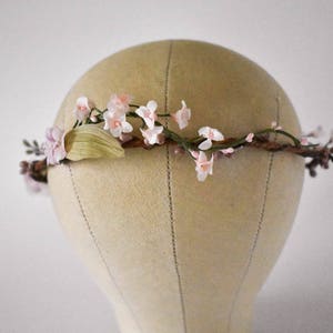 Puede incluir: Una corona floral con delicadas flores rosas y blancas, hojas verdes y peque&ntilde;os capullos marrones. Las flores est&aacute;n dispuestas sobre una base marr&oacute;n en forma de ramita. La corona se exhibe en una cabeza de maniqu&iacute;.