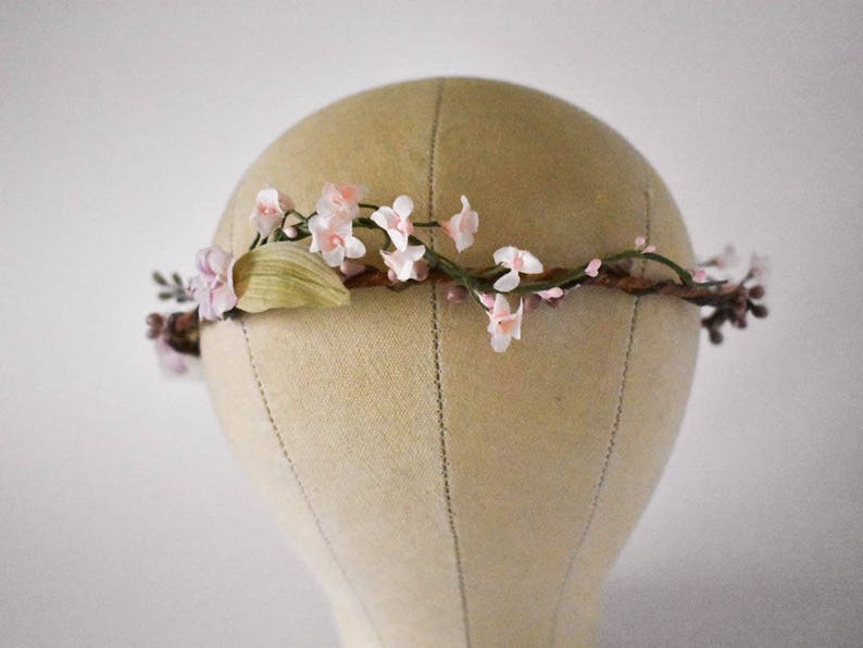 Puede incluir: Una corona floral con delicadas flores rosas y blancas, hojas verdes y peque&ntilde;os capullos marrones. Las flores est&aacute;n dispuestas sobre una base marr&oacute;n en forma de ramita. La corona se exhibe en una cabeza de maniqu&iacute;.