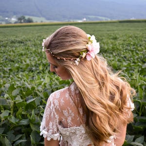 Puede incluir: Una mujer con largo cabello rubio ondulado, adornada con una corona floral con flores rosas y blancas. Lleva una blusa de encaje blanco, frente a un campo de plantas verdes. Se ve una cordillera al fondo.