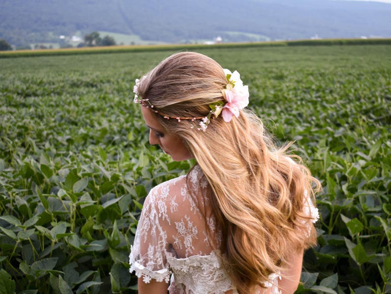 Puede incluir: Una mujer con largo cabello rubio ondulado, adornada con una corona floral con flores rosas y blancas. Lleva una blusa de encaje blanco, frente a un campo de plantas verdes. Se ve una cordillera al fondo.