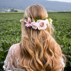 Puede incluir: Una mujer con largo cabello rubio ondulado, adornada con una corona floral de flores blancas y rosas. De espaldas a la c&aacute;mara, con un campo verde de fondo. Lleva una blusa de encaje blanco.
