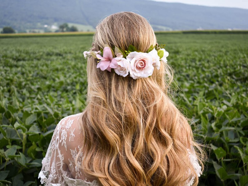 Puede incluir: Una mujer con largo cabello rubio ondulado, adornada con una corona floral de flores blancas y rosas. De espaldas a la c&aacute;mara, con un campo verde de fondo. Lleva una blusa de encaje blanco.