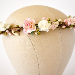 Op de afbeelding: Een bloemenkrans met roze en witte papieren rozen, groene knoppen en kleine roze bessen. De bloemen zijn bevestigd aan een bruine, takachtige basis. De krans wordt getoond op een mannequinhoofd.