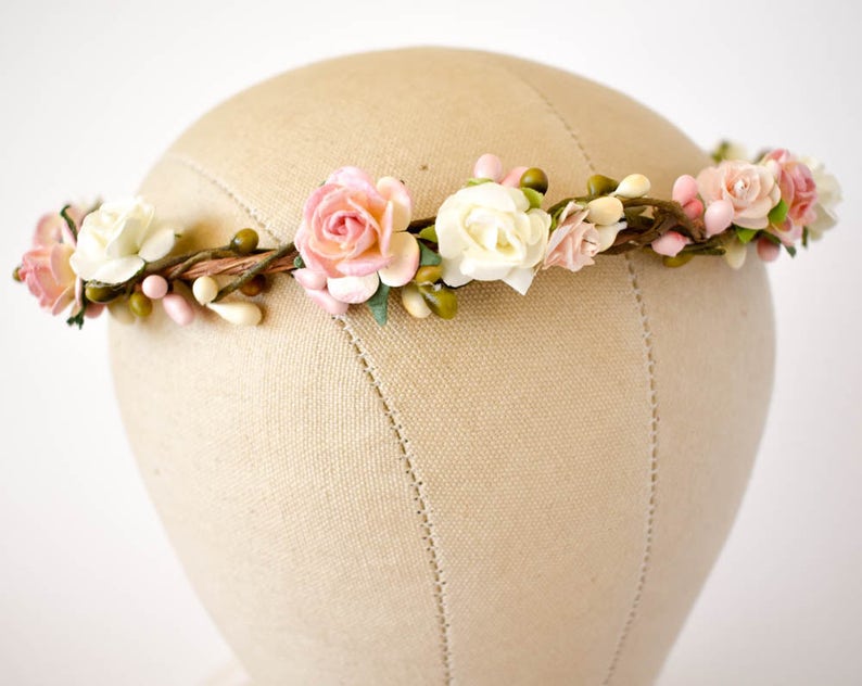 Op de afbeelding: Een bloemenkrans met roze en witte papieren rozen, groene knoppen en kleine roze bessen. De bloemen zijn bevestigd aan een bruine, takachtige basis. De krans wordt getoond op een mannequinhoofd.