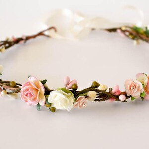 Op de afbeelding: Een bloemenkrans met kunstbloemen in een cirkelvormige opstelling. Het heeft witte, lichtroze en perzikrozen, kleine groene knoppen en roze bessen. De basis is gemaakt van bruine twijgen en een cr&egrave;mekleurig lint is bevestigd.
