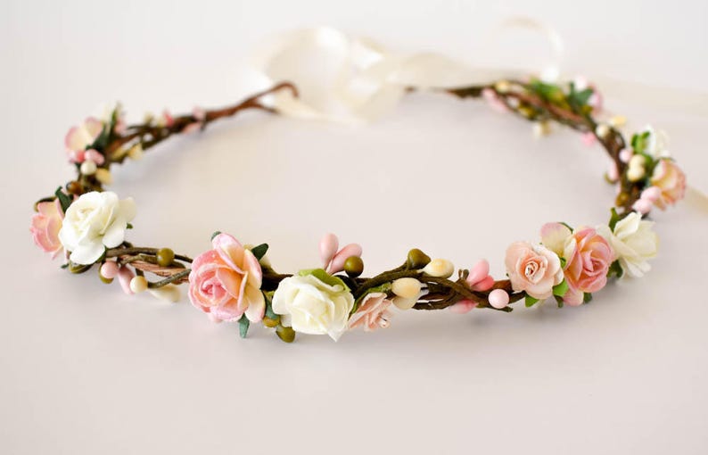 Op de afbeelding: Een bloemenkrans met kunstbloemen in een cirkelvormige opstelling. Het heeft witte, lichtroze en perzikrozen, kleine groene knoppen en roze bessen. De basis is gemaakt van bruine twijgen en een cr&egrave;mekleurig lint is bevestigd.