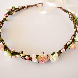 Op de afbeelding: Een bloemenkrans met een bruine wijnstokbasis versierd met witte, roze en perzikbloemen en kleine witte bessen. De kroon is vastgebonden met een cr&egrave;mekleurig lint, perfect voor bruiloften of speciale gelegenheden.