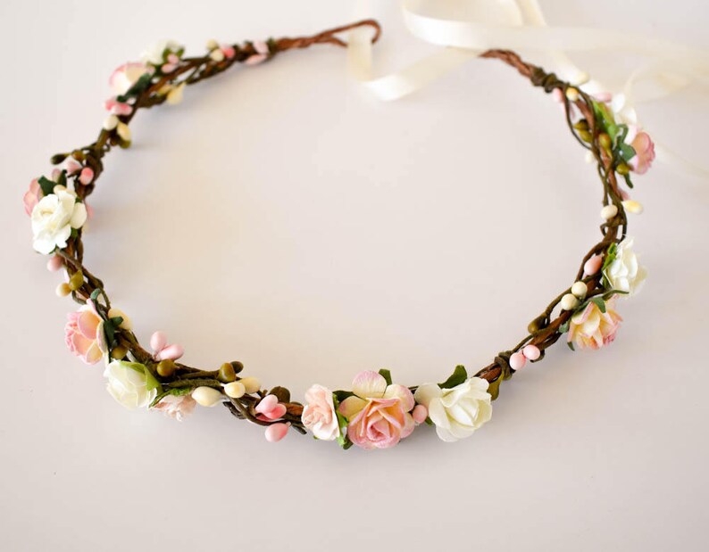 Op de afbeelding: Een bloemenkrans met een bruine wijnstokbasis versierd met witte, roze en perzikbloemen en kleine witte bessen. De kroon is vastgebonden met een cr&egrave;mekleurig lint, perfect voor bruiloften of speciale gelegenheden.