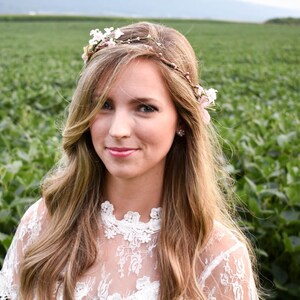 Puede incluir: Una mujer con cabello rubio largo y ondulado, con una corona de flores y una blusa de encaje blanco. El fondo es un campo de plantas verdes. La mujer sonr&iacute;e a la c&aacute;mara.