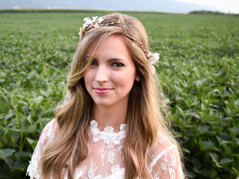 Puede incluir: Una mujer con cabello rubio largo y ondulado, con una corona de flores y una blusa de encaje blanco. El fondo es un campo de plantas verdes. La mujer sonr&iacute;e a la c&aacute;mara.
