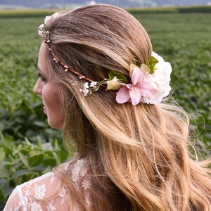 Puede incluir: Una mujer con cabello largo y ondulado rubio, adornada con una corona floral y un racimo de flores. La corona est&aacute; hecha de peque&ntilde;as flores blancas y ramitas marrones. Las flores son rosas y blancas, con hojas verdes. Lleva un top de encaje blanco.