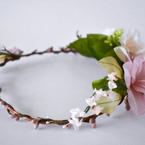 Puede incluir: Una corona floral con un dise&ntilde;o delicado. La corona est&aacute; hecha de vides marrones y retorcidas adornadas con peque&ntilde;os capullos rosados y racimos de flores blancas y rosas. Tambi&eacute;n hay grandes flores rosas y blancas con estambres amarillos.