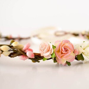 Op de afbeelding: Een bloemenkrans met roze en cr&egrave;mekleurige rozen, knoppen en groen. De bloemen zijn gerangschikt op een bruine, takachtige basis, met een zacht, wit lint dat zich naar achteren uitstrekt. Deze delicate hoofdtooi is geschikt voor bruiloften of speciale gelegenheden.