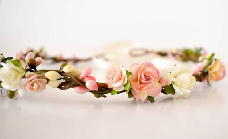 Op de afbeelding: Een bloemenkrans met roze en cr&egrave;mekleurige rozen, knoppen en groen. De bloemen zijn gerangschikt op een bruine, takachtige basis, met een zacht, wit lint dat zich naar achteren uitstrekt. Deze delicate hoofdtooi is geschikt voor bruiloften of speciale gelegenheden.