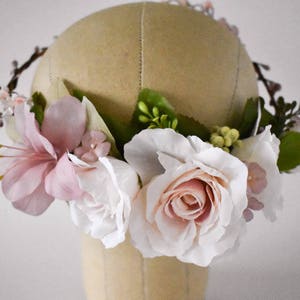 Puede incluir: Una corona floral con rosas blancas y rosadas artificiales, lirios rosas y peque&ntilde;as flores rosas. Hojas verdes y ramitas marrones se intercalan entre las flores. La corona se exhibe en una cabeza de maniqu&iacute; beige.