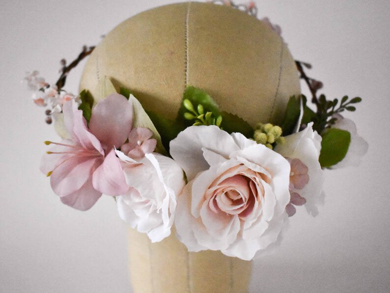 Puede incluir: Una corona floral con rosas blancas y rosadas artificiales, lirios rosas y peque&ntilde;as flores rosas. Hojas verdes y ramitas marrones se intercalan entre las flores. La corona se exhibe en una cabeza de maniqu&iacute; beige.
