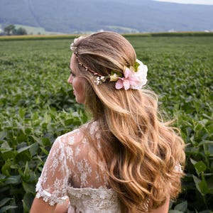 Puede incluir: Una mujer con cabello largo y ondulado rubio, adornado con una corona floral y una flor rosa. Viste un vestido de encaje blanco, de pie en un campo de plantas verdes con una cordillera al fondo. La imagen captura una est&eacute;tica natural y rom&aacute;ntica.